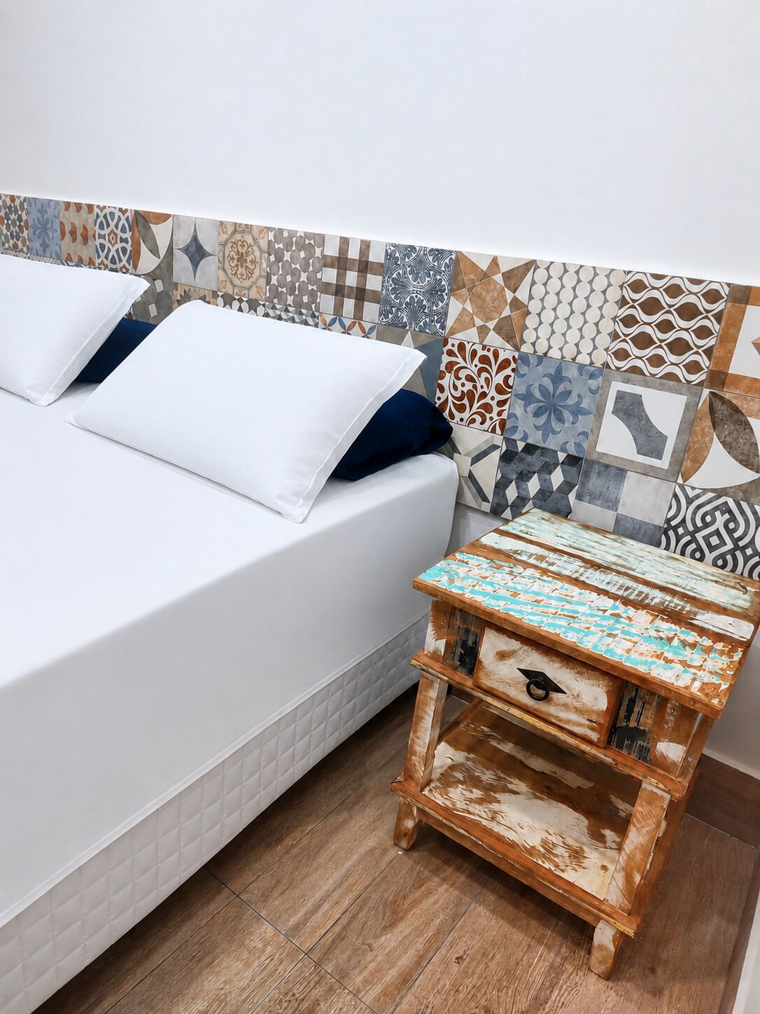 A bed with white bedding, a wooden nightstand with a patterned top, and a tiled wall with various designs.