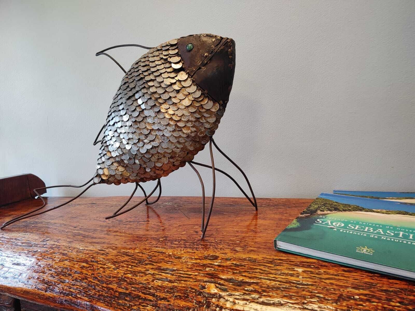 Une sculpture décorative représentant un poisson, réalisée à partir de pièces de monnaie, est posée sur une surface en bois, à côté d’un livre intitulé « SÃO SEBASTIÃO ».