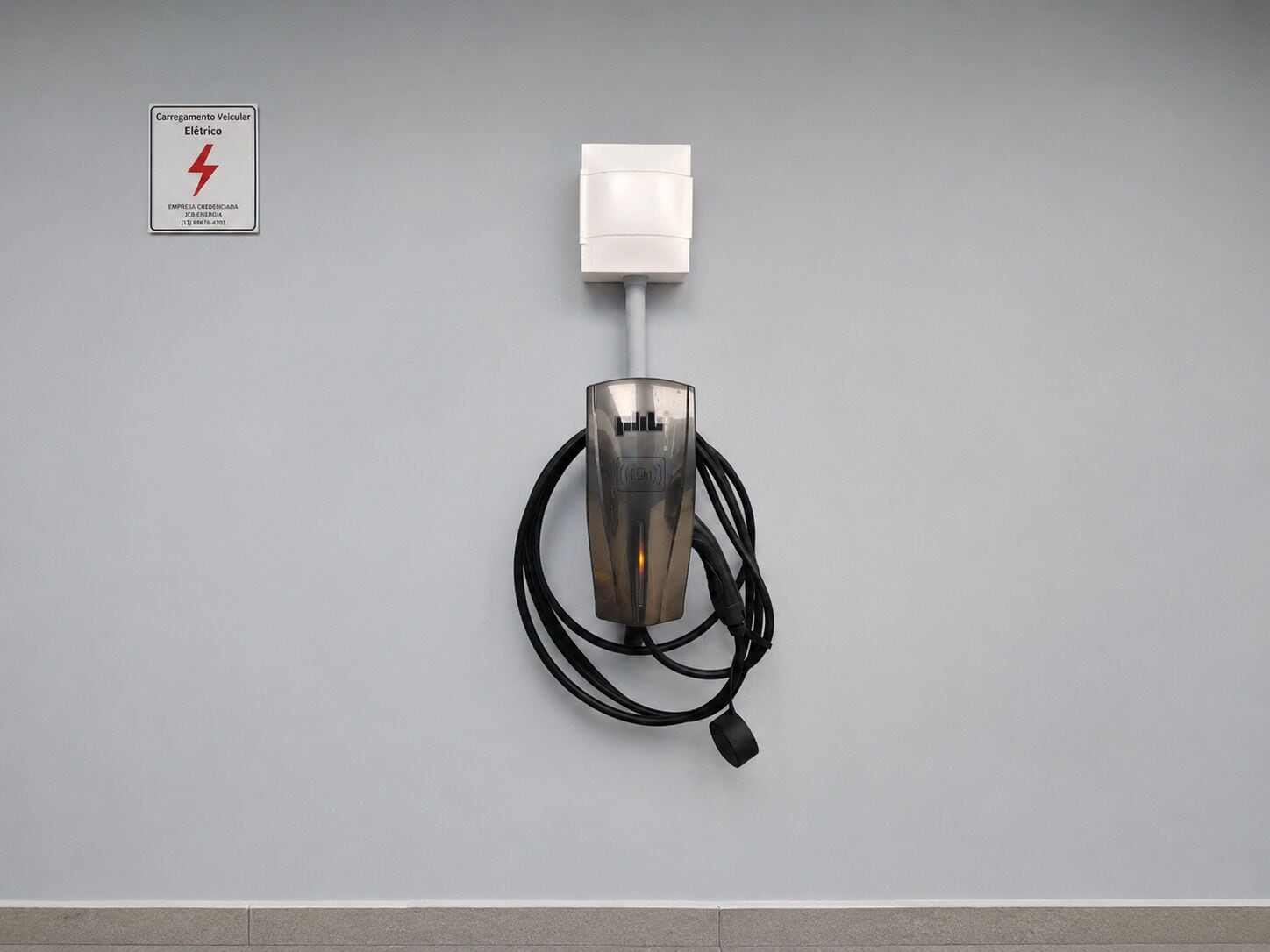 Uma estação de carregamento para veículos elétricos fixada na parede, com um cabo e uma placa indicando ser um ponto de carregamento elétrico.