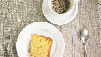 A slice of cake on a white plate, a cup of coffee, a fork, a knife, and a spoon on a textured tablecloth.