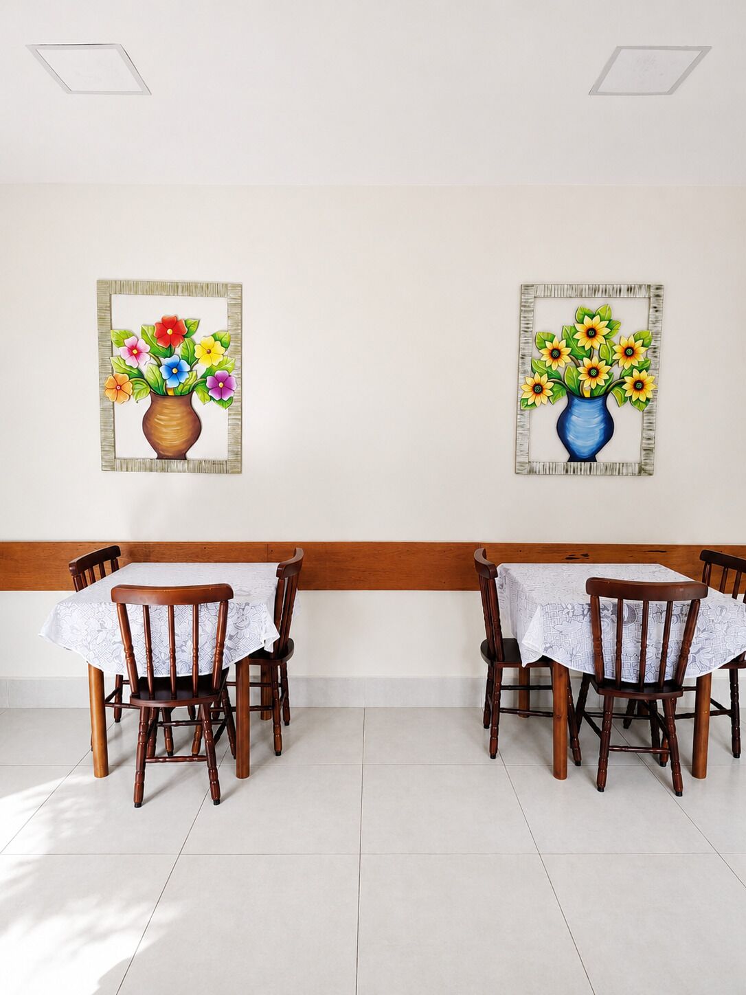 Un'area pranzo con una panca e sedie di legno, un pavimento in piastrelle bianche e due quadri appesi raffiguranti vasi con fiori.