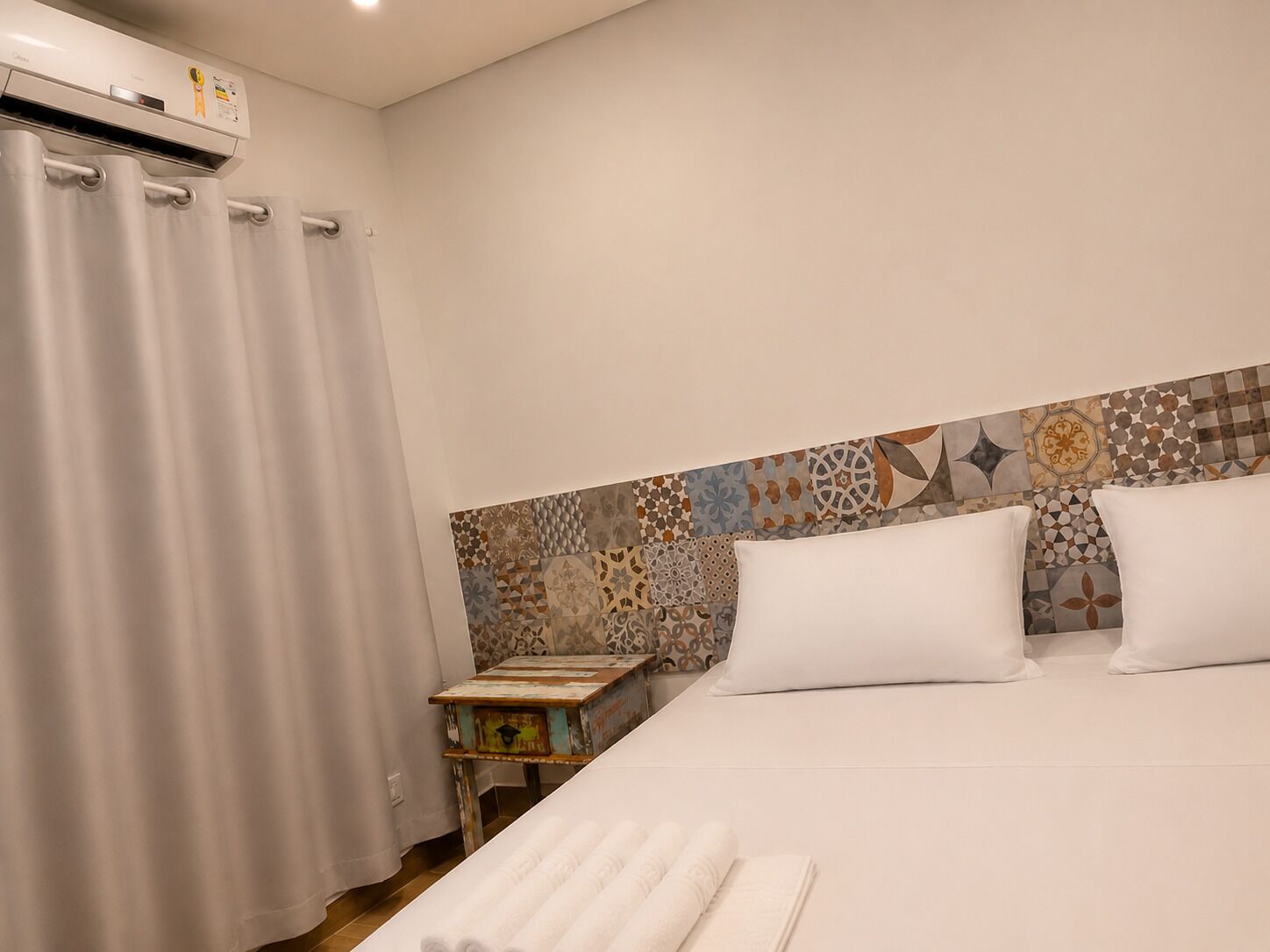 Hotelkamer met een bed, witte kussens, een gestreept hoofdeinde, een kleine tafel met een lamp, en een gordijn.