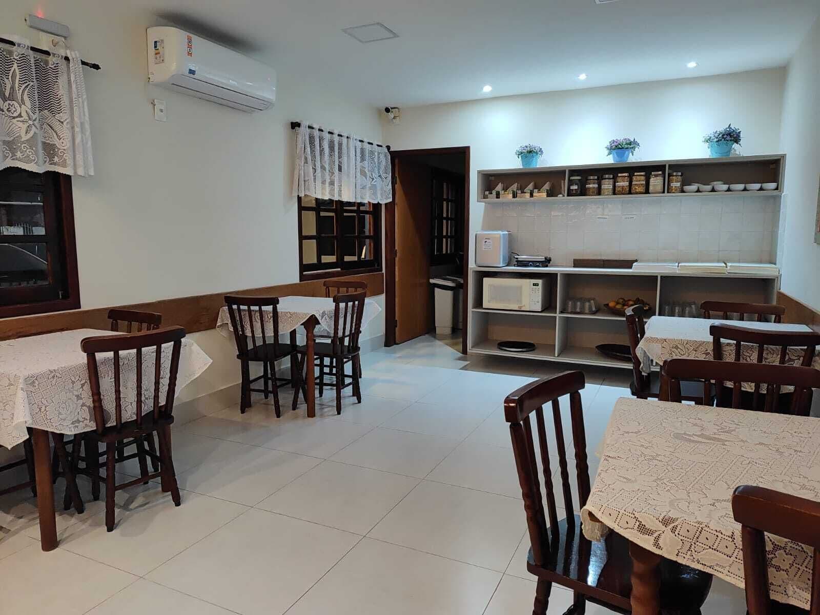 A dining area with wooden chairs and tables, a kitchen area with shelves and appliances, and a window with lace curtains.