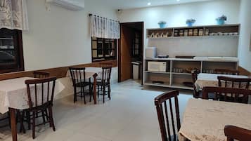 A dining area with wooden chairs and tables, a kitchen area with shelves and appliances, and a window with lace curtains.