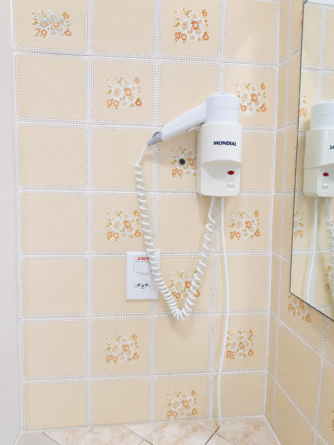 A bathroom with a hairdryer mounted on the wall, a power outlet, and a mirror reflecting the bathroom.