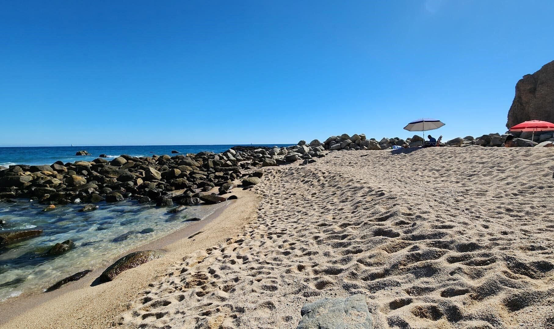 Una spiaggia sabbiosa con acque cristalline, grandi rocce e un ombrellone rosso.