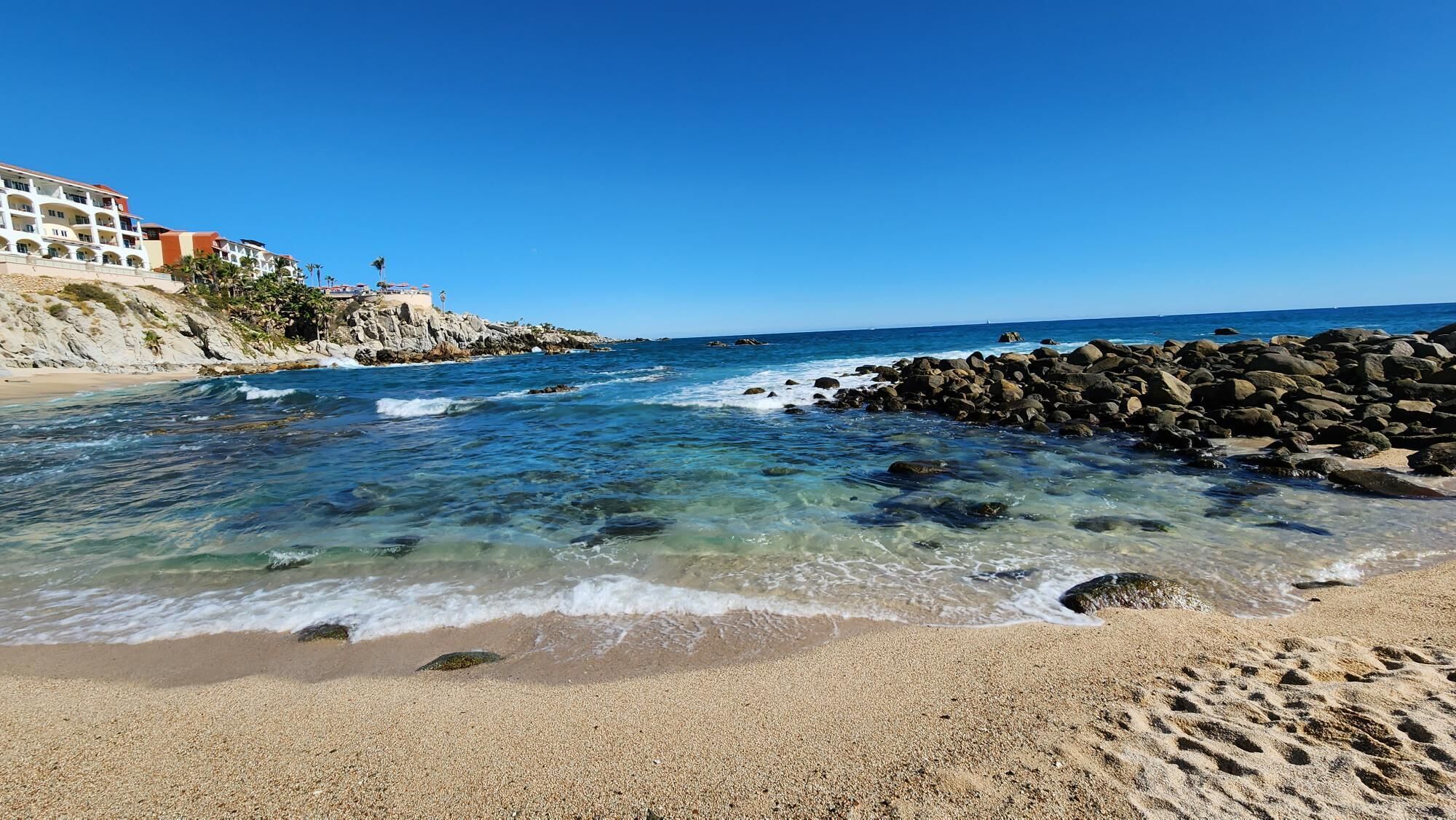 Una playa con agua cristalina, arena pedregosa y un rompeolas rocoso.