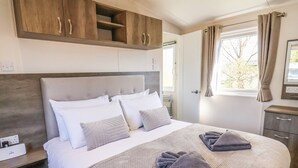 A neatly made bed with a grey blanket and pillows, a wooden headboard, and a window with curtains. - Honeysuckle Lodge (Saltburn-by-the-Sea)