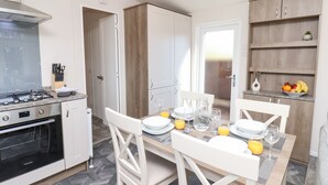 A modern kitchen with white cabinetry, a gas stove, and a built-in oven. There is a dining area with a table set for two, white chairs, and a fruit bowl on the sideboard. - Honeysuckle Lodge (Saltburn-by-the-Sea)