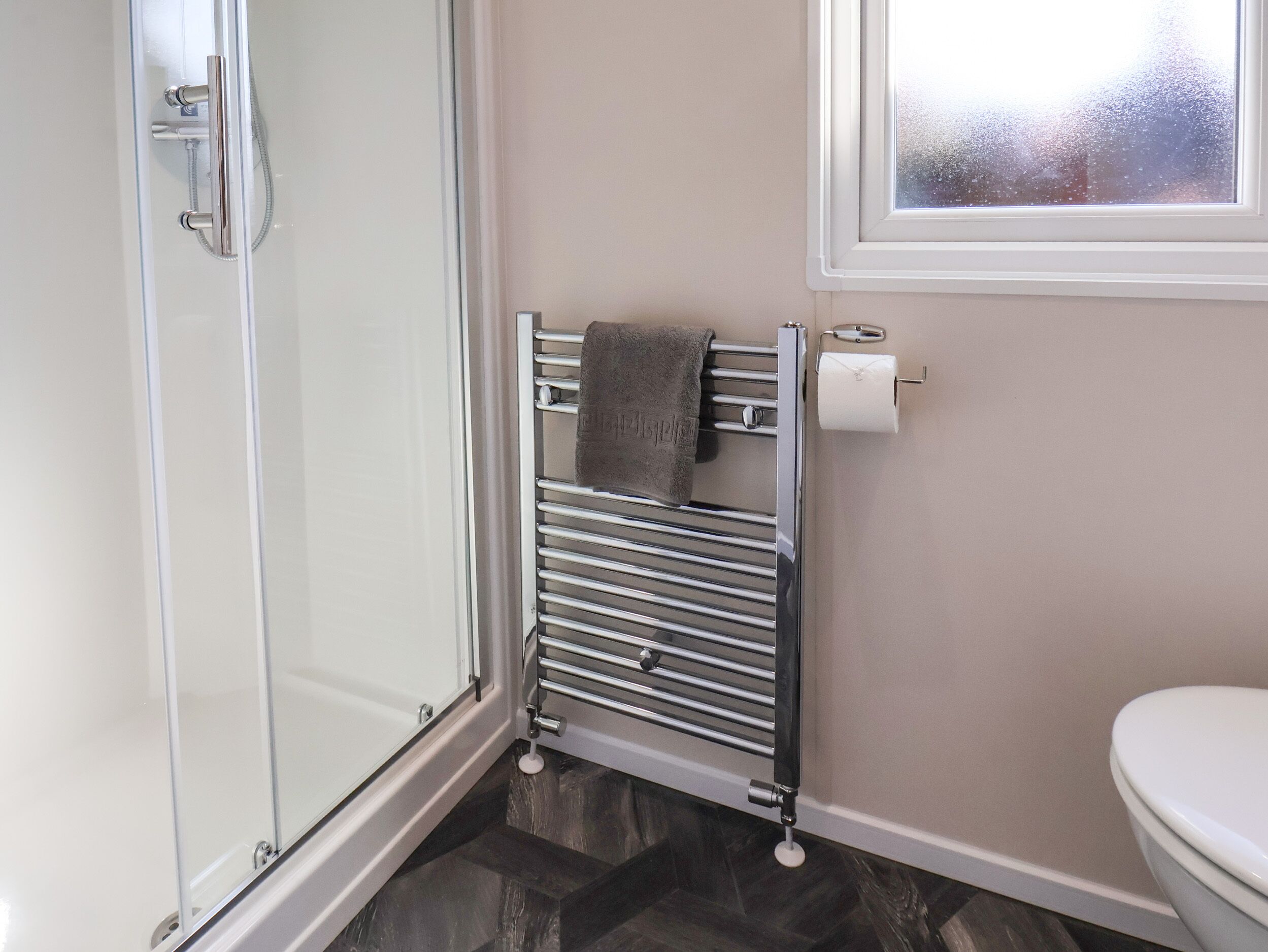 A bathroom with a glass shower door, a towel rack with a towel, a toilet, and a window with frosted glass.