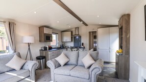 A modern living room with a grey sofa set, a coffee table with croissants, and a kitchen area with a microwave and stove. - Honeysuckle Lodge (Saltburn-by-the-Sea)
