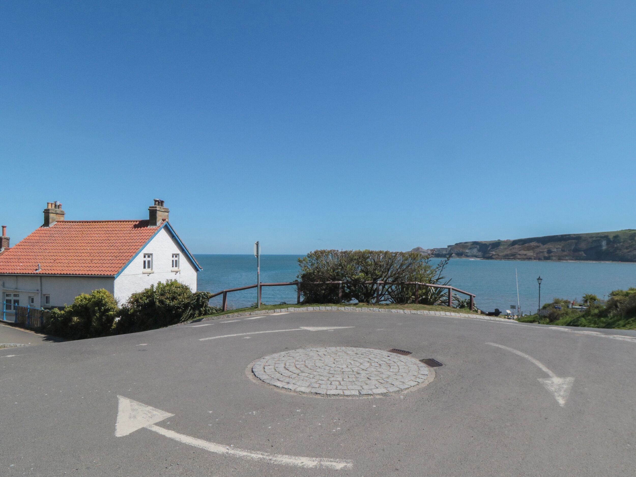 A coastal road with a roundabout, a white building with a red roof, and a view of the sea.