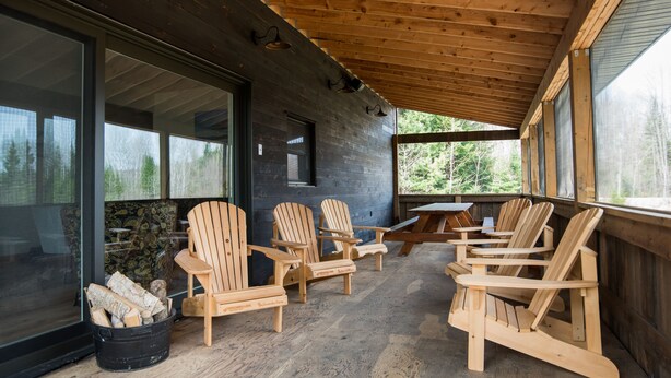 A covered porch with wooden chairs and a picnic table, large windows, and a black bucket with logs. - Country Cottage on 80 acres (Burk's Falls)