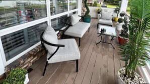 Een overdekt terras buiten met houten vloer, witte zachte zitplaatsen en potten met planten.