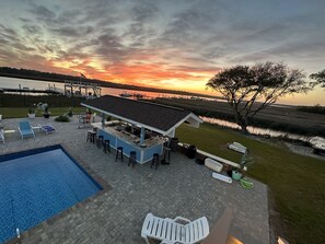 Un espace piscine doté de chaises longues et d’un bar, offrant une vue sur un plan d’eau et un magnifique coucher de soleil.