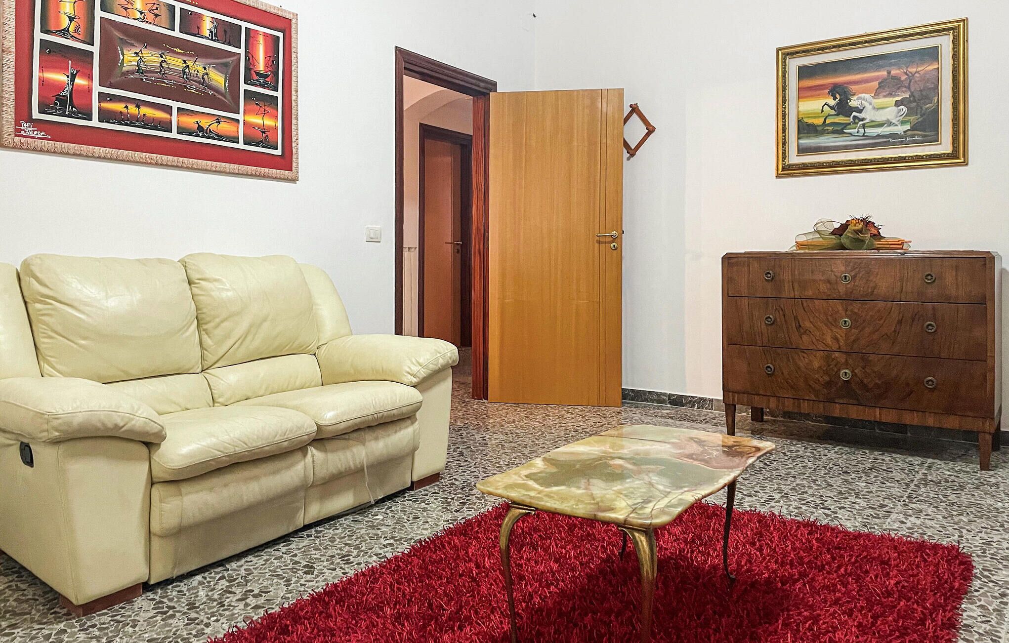 Un salon avec un canapé en cuir beige, une petite table basse en verre, un tapis rouge à longues mèches, une commode en bois et une porte.