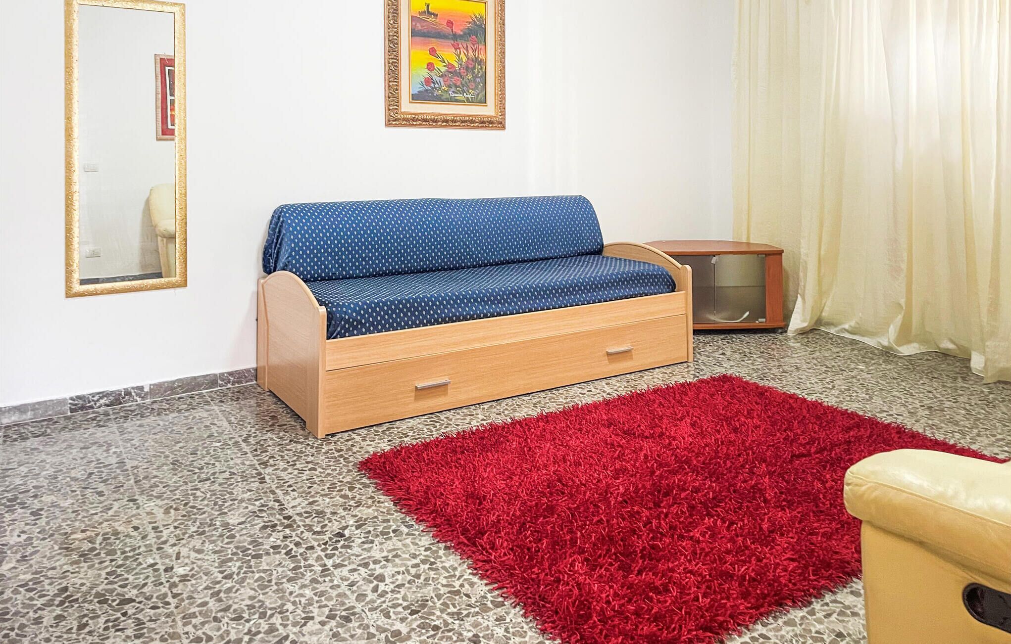Una habitación con un sofá azul, un tapete rojo, un armario de madera y un espejo en la pared.