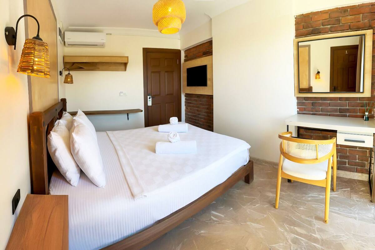 A bedroom with a bed, a desk, a chair, a mirror, and a hanging light fixture.