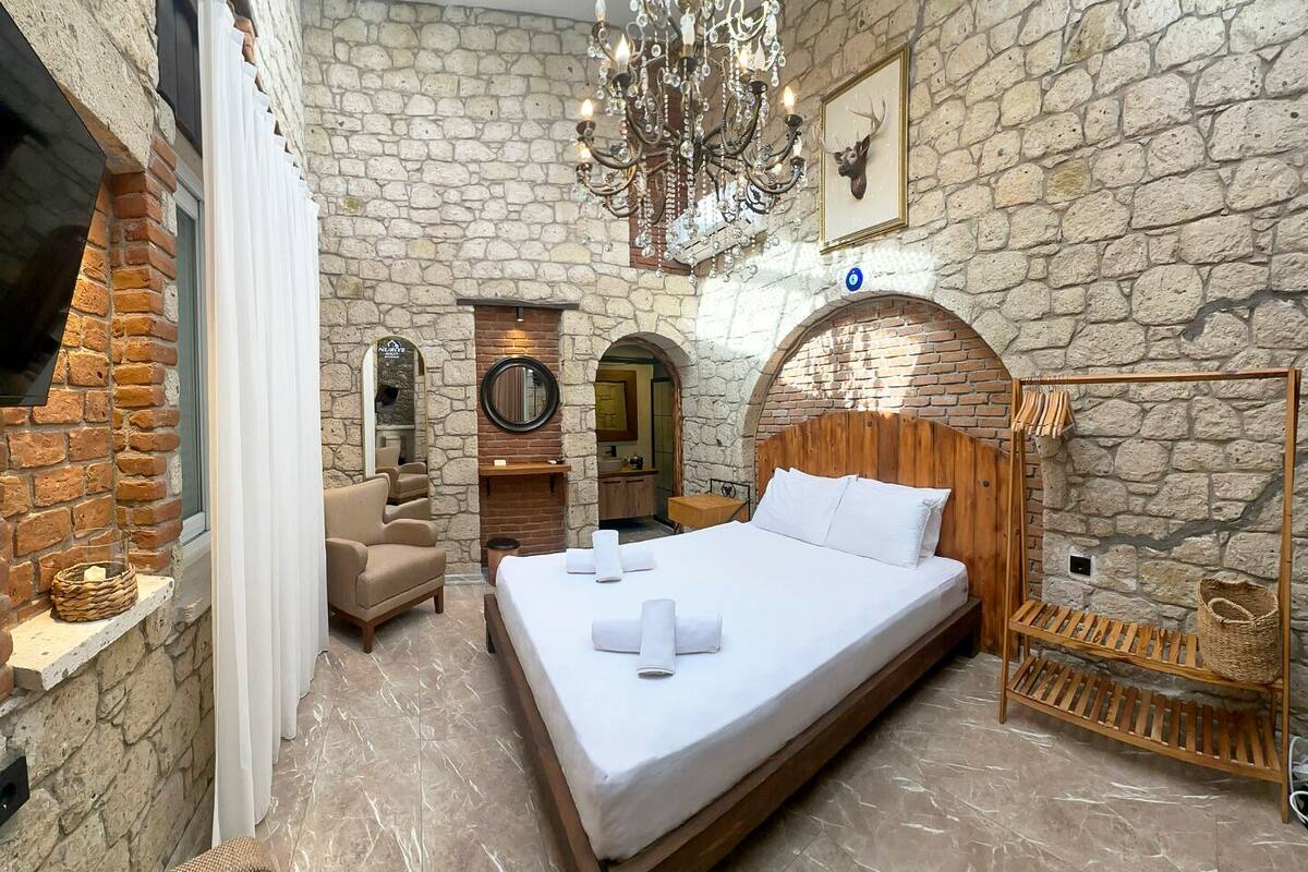 A bedroom with a stone wall, a wooden headboard, a chandelier, a bed with white linens, a chair, and a wooden shelf.