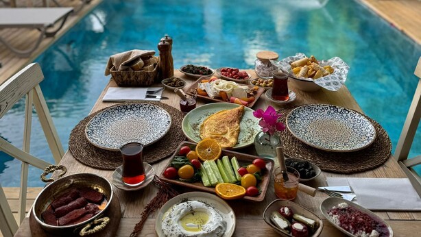 A poolside dining setup with a variety of foods and drinks, a view of a building with a sign, and outdoor furniture. - Nuriye Alacati (Cesme)