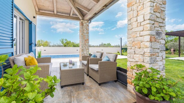 Un espace extérieur abrité, aménagé avec des meubles en osier, des piliers en pierre et offrant une vue sur une piscine.