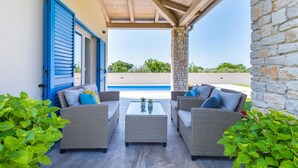 Un espace extérieur aménagé, abrité et doté de mobilier en osier, d’une table en verre et offrant une vue sur une piscine.