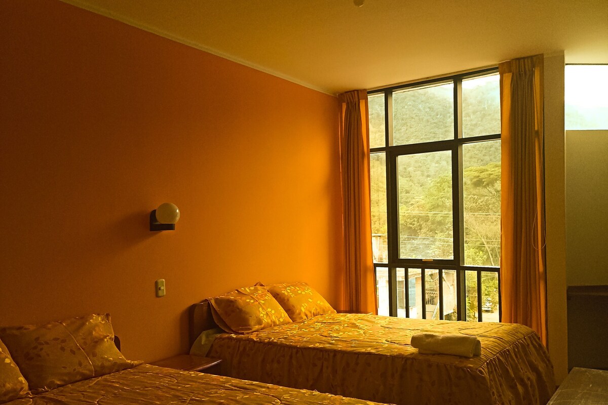 A room with two beds, a window with curtains, a small table, and a wall-mounted light fixture.