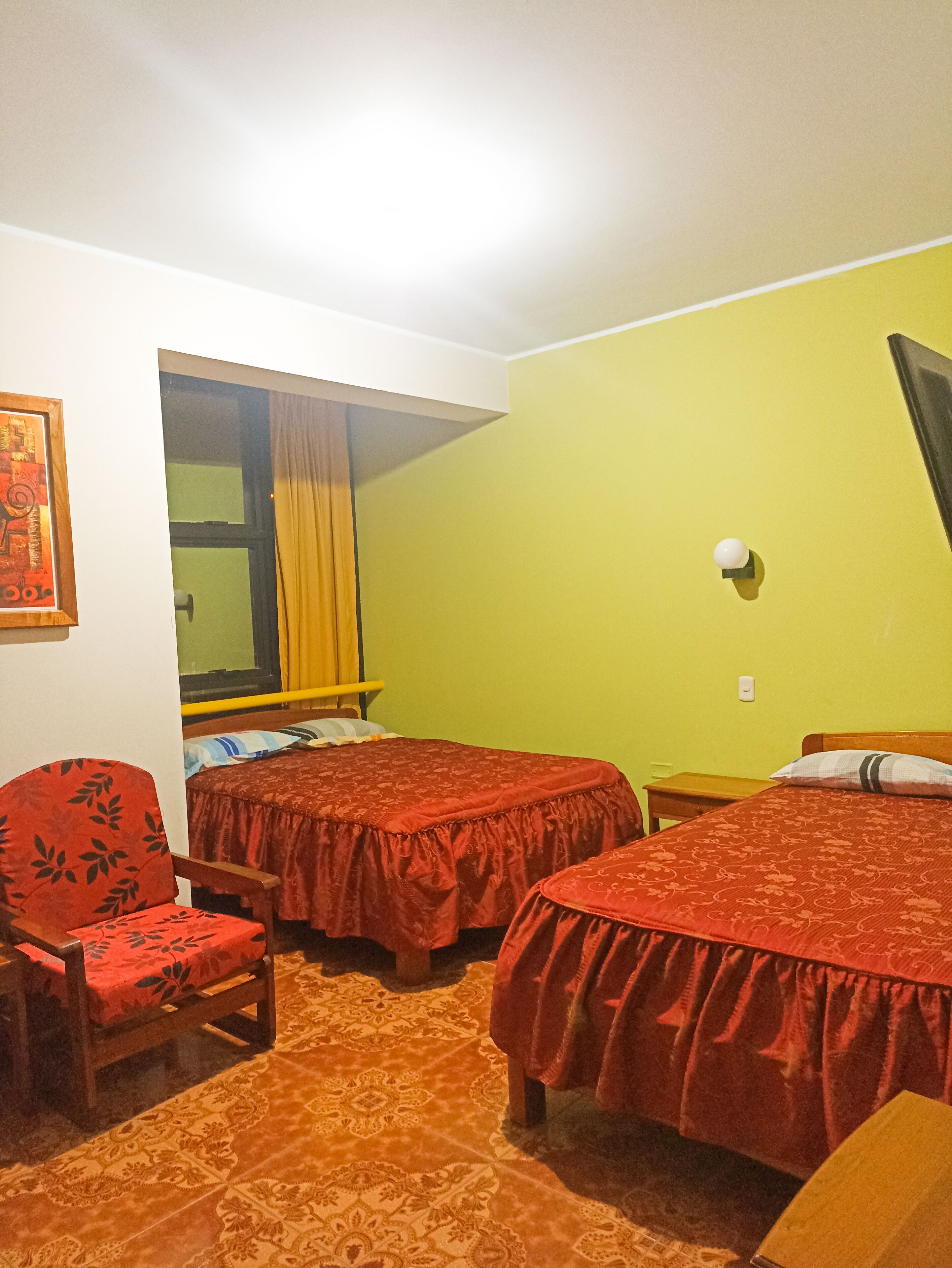 A room with two beds, a chair, a window with curtains, and a wall-mounted light.
