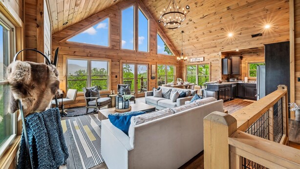 A spacious wooden cabin with a high ceiling, large windows, and a cozy living area with a sofa and dining table. - Mtn Cabin · Hot Tub · Game Room · Fire Pit (Sevierville)