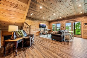 A cozy cabin interior with wooden walls, a TV, leather sofas, and a large window offering a view of greenery. - Views for miles · Hot Tub · Game Room · Fire Pit (Sevierville)