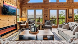 A cozy cabin interior with wooden walls, a fireplace, comfortable seating, and large windows offering scenic views. - Mtn Cabin · Hot Tub · Game Room · Fire Pit (Sevierville)