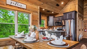 A wooden interior with a dining table set for four, a kitchen with modern appliances, and a view of greenery outside. - Mtn Cabin · Hot Tub · Game Room · Fire Pit (Sevierville)