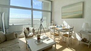 A room with a large window, a white table, a white chair, a wicker chair, a tripod, and a painting on the wall. - Les Pieds dans l'Eau, bay view for 6 people (Le Crotoy)