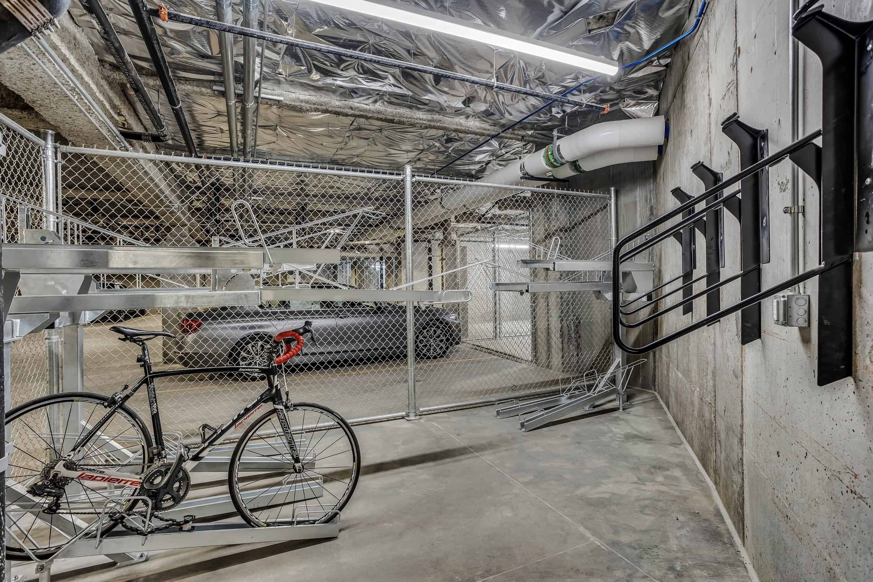 Una bicicleta fijada a un soporte metálico en un área de estacionamiento techada, con cerramiento de malla metálica, piso de concreto y techo industrial con tuberías a la vista.