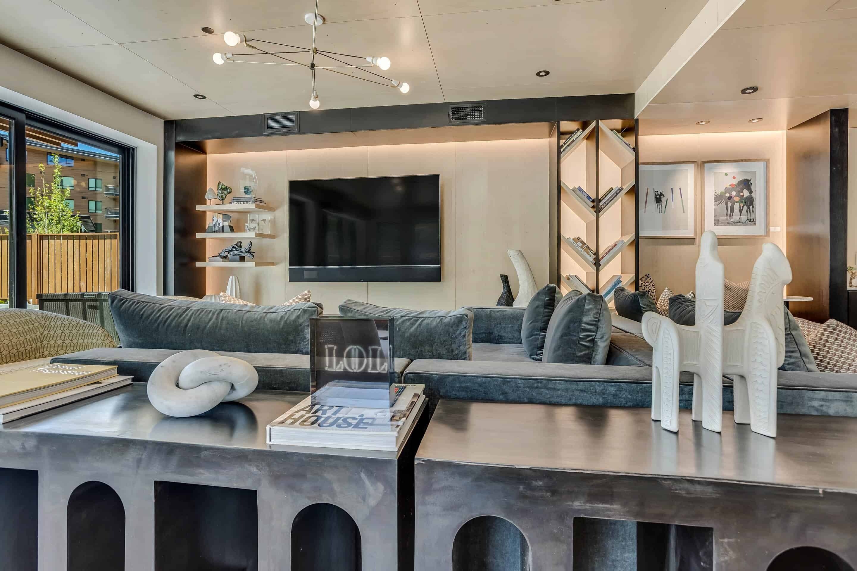 Ein modernes Wohnzimmer mit einem großen Flachbildfernseher, grauen Sofas, einem Couchtisch mit Büchern und einer Skulptur sowie einem Bücherregal mit gerahmten Bildern.