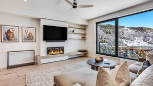 A modern living room with a fireplace, a flat-screen TV, and a large window offering a view of a snowy landscape. - Electric Pass Lodge (Snowmass Village)