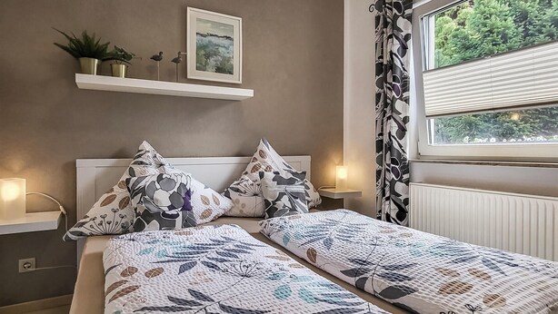 A bedroom with two single beds, a bedside table with a lamp, a shelf with decorative items, and a window with curtains. - 1 bedroom cozy apartment in Börgerende (Börgerende)