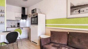 A compact kitchen with white cabinets, a microwave, and a stove. There is a dining table with chairs, a small round table, and a brown sofa. A framed picture hangs on the wall. - 1 bedroom cozy apartment in Börgerende (Börgerende)
