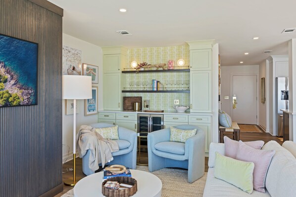 A living room with a sofa, two armchairs, a coffee table, and a kitchen area with light green cabinetry and a countertop.