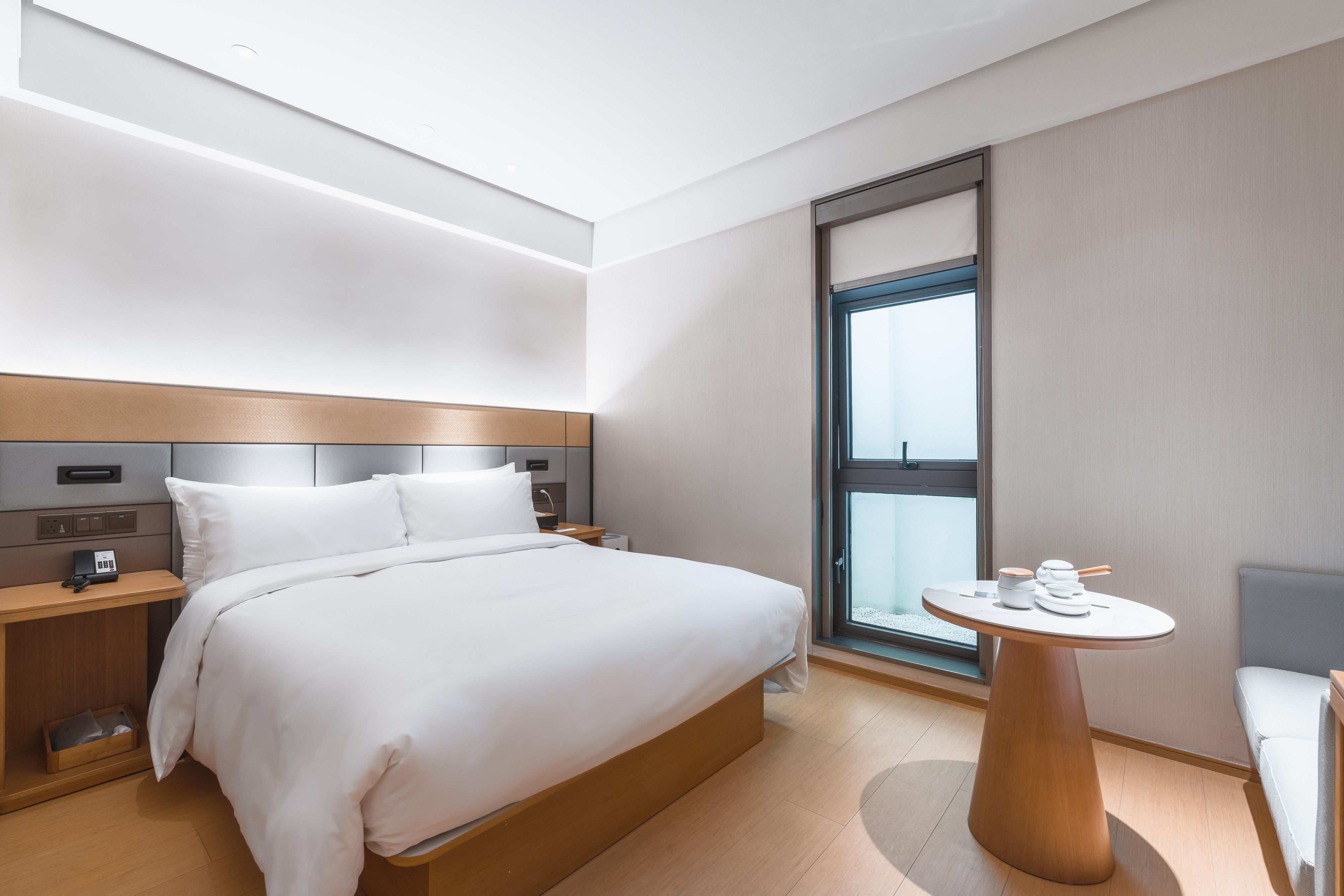 A modern hotel room with a large bed, bedside tables, a small round table with a lamp, and a window with a view.
