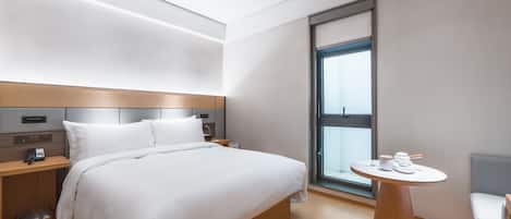 A modern hotel room with a large bed, bedside tables, a small round table with a lamp, and a window with a view.