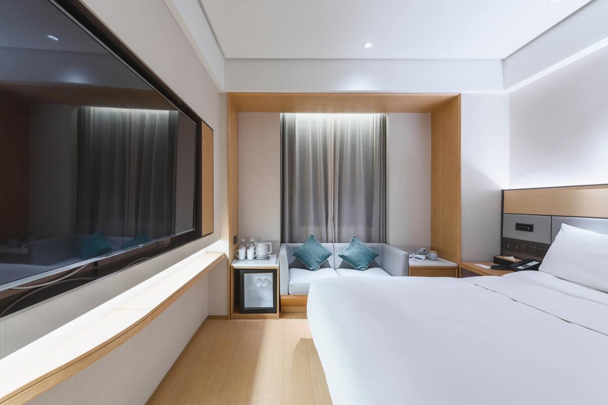 A modern hotel room with a large bed, a sofa, a small table with a phone and a cup, and a window with curtains.