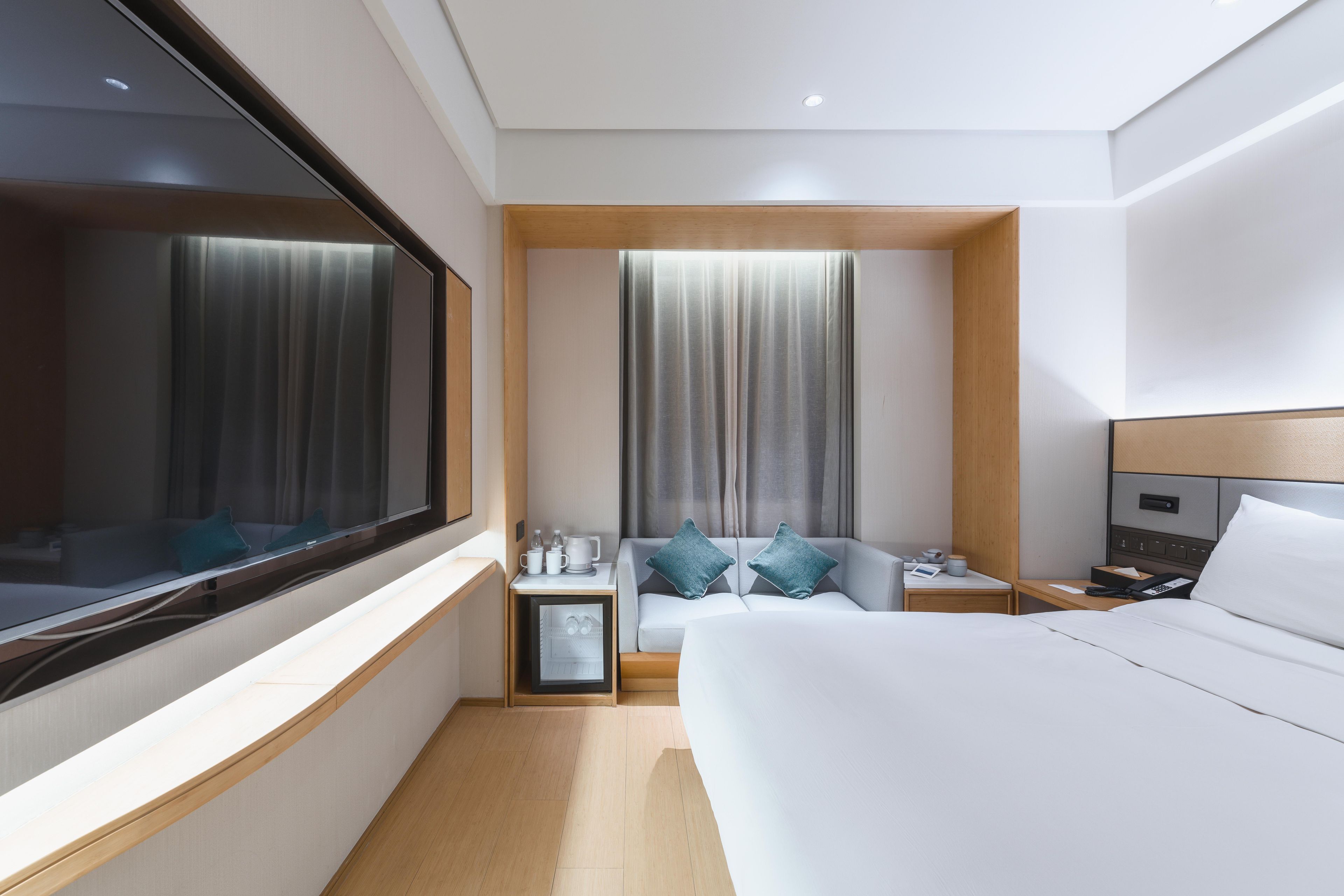 A modern hotel room with a large bed, a sofa, a small table with a phone and a cup, and a window with curtains.