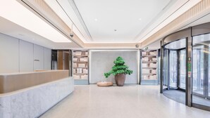 Un hall d’hôtel moderne avec un comptoir d’accueil, une grande plante en pot et un ascenseur en verre.