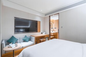 Quarto de hotel com cama, área de estar com mesa, televisão, escrivaninha com espelho e banheiro com pia e chuveiro.
