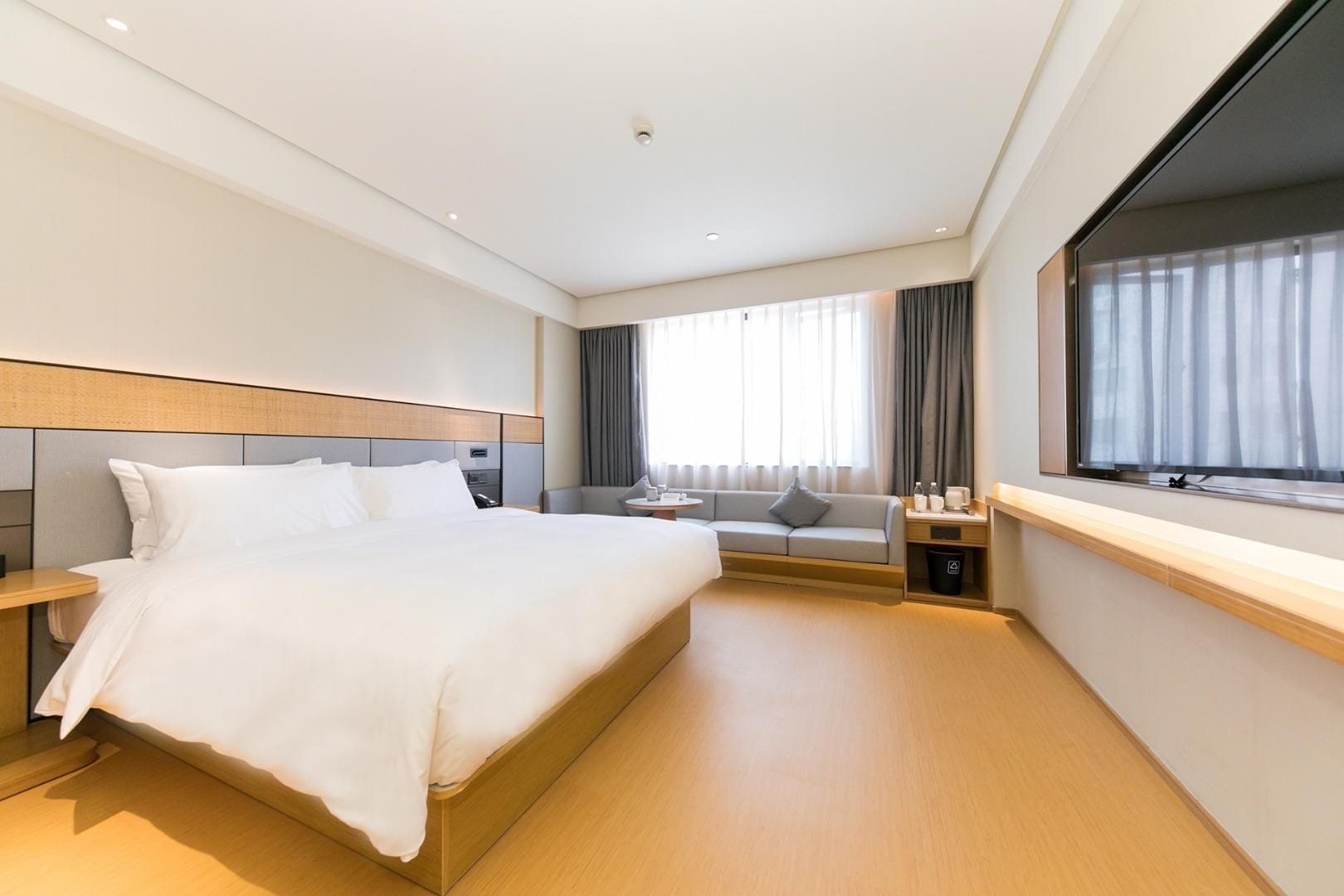 A modern hotel room with a large bed, a sofa, a small table, and a window with curtains.