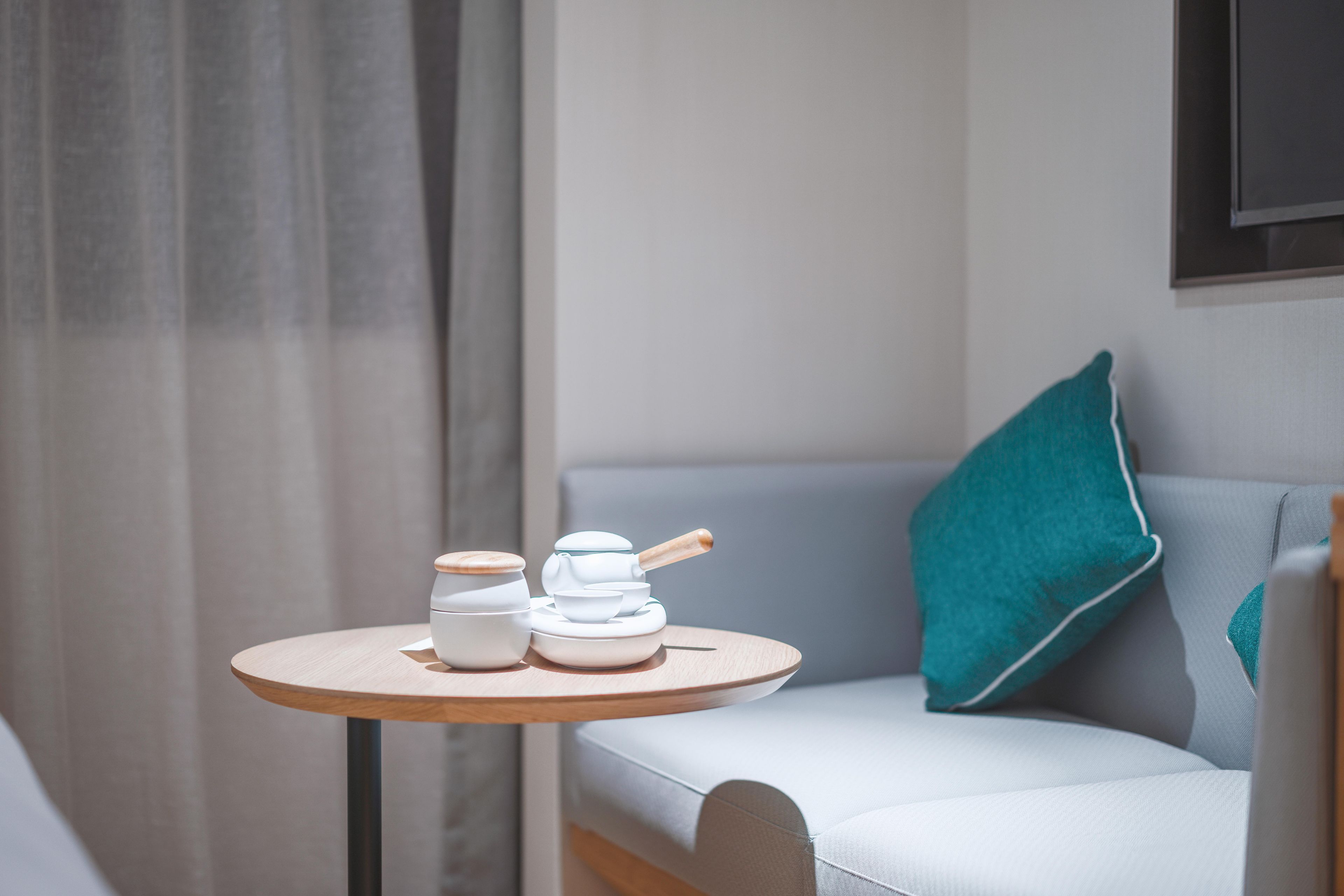 A small round table with two white ceramic containers and a wooden spoon, placed on a light-colored sofa with a teal pillow.
