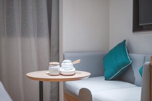 Une petite table ronde sur laquelle sont posés deux récipients en céramique blanche et une cuillère en bois, le tout placé sur un canapé de couleur claire agrémenté d’un coussin turquoise.