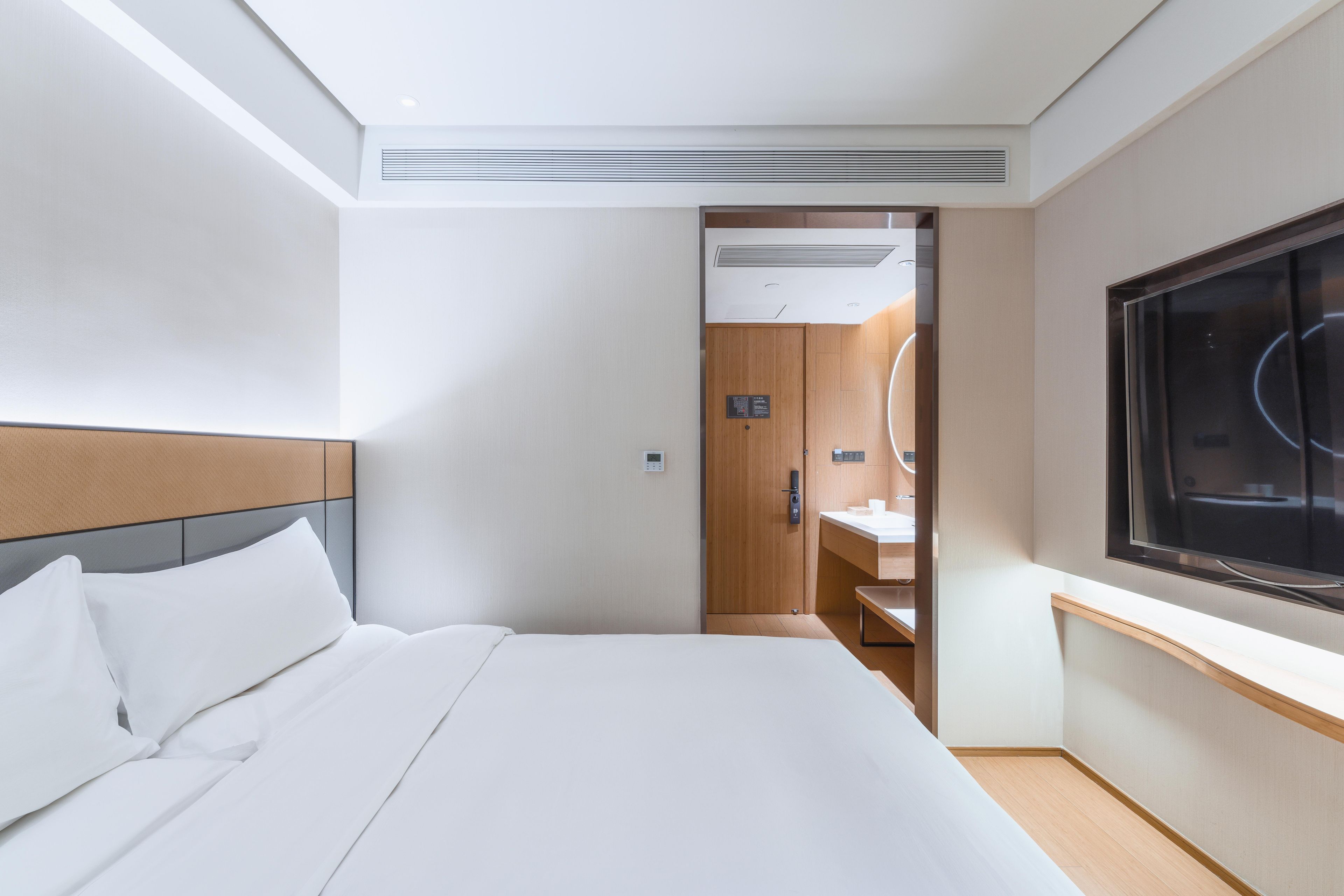 A hotel room with a bed, a large window, a wooden door, and a wall-mounted TV.