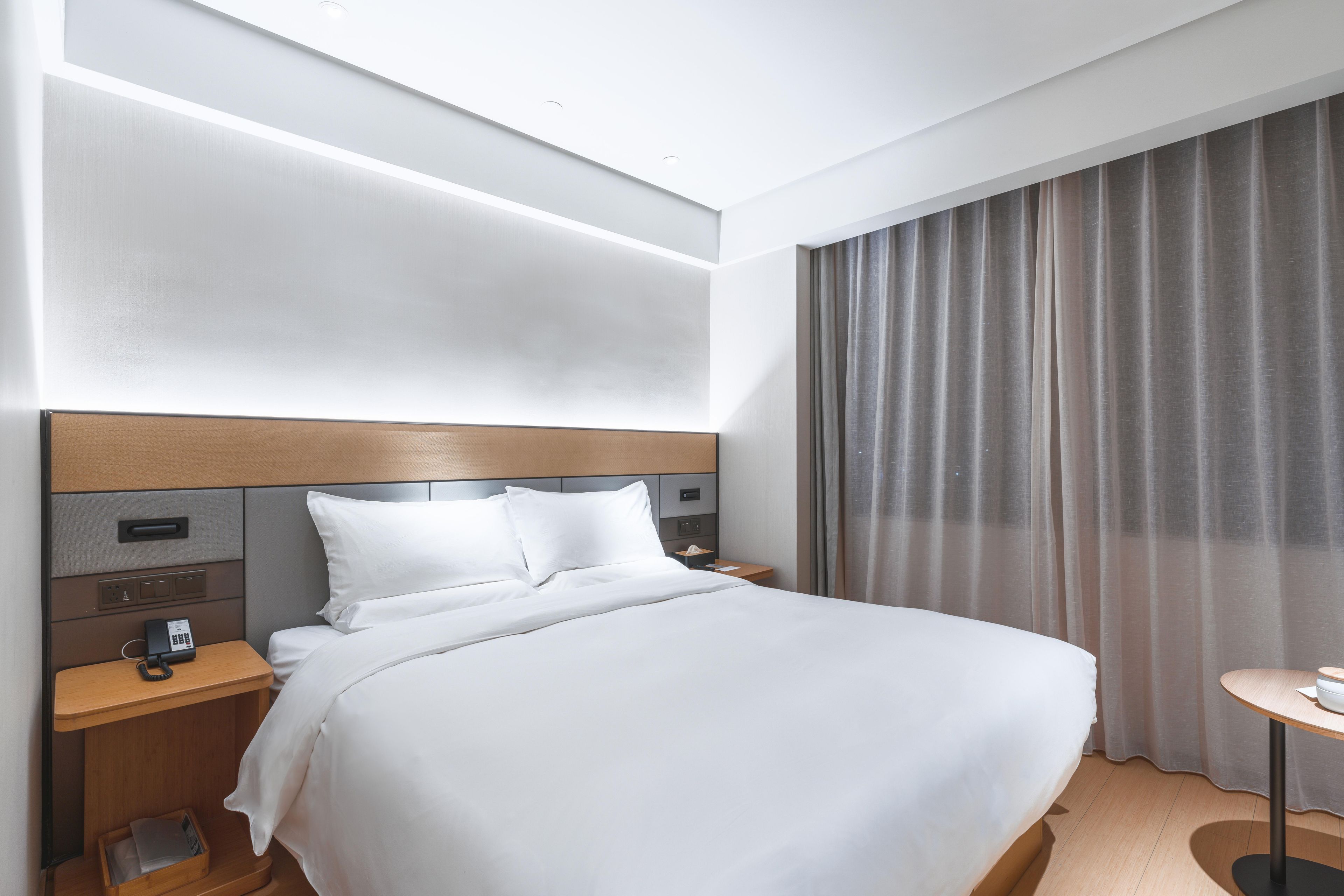 Ein Hotelzimmer mit einem großen Bett, einem Nachttisch mit einer Lampe, einem weiteren Nachttisch mit einem Telefon und einem Fenster mit Vorhängen.